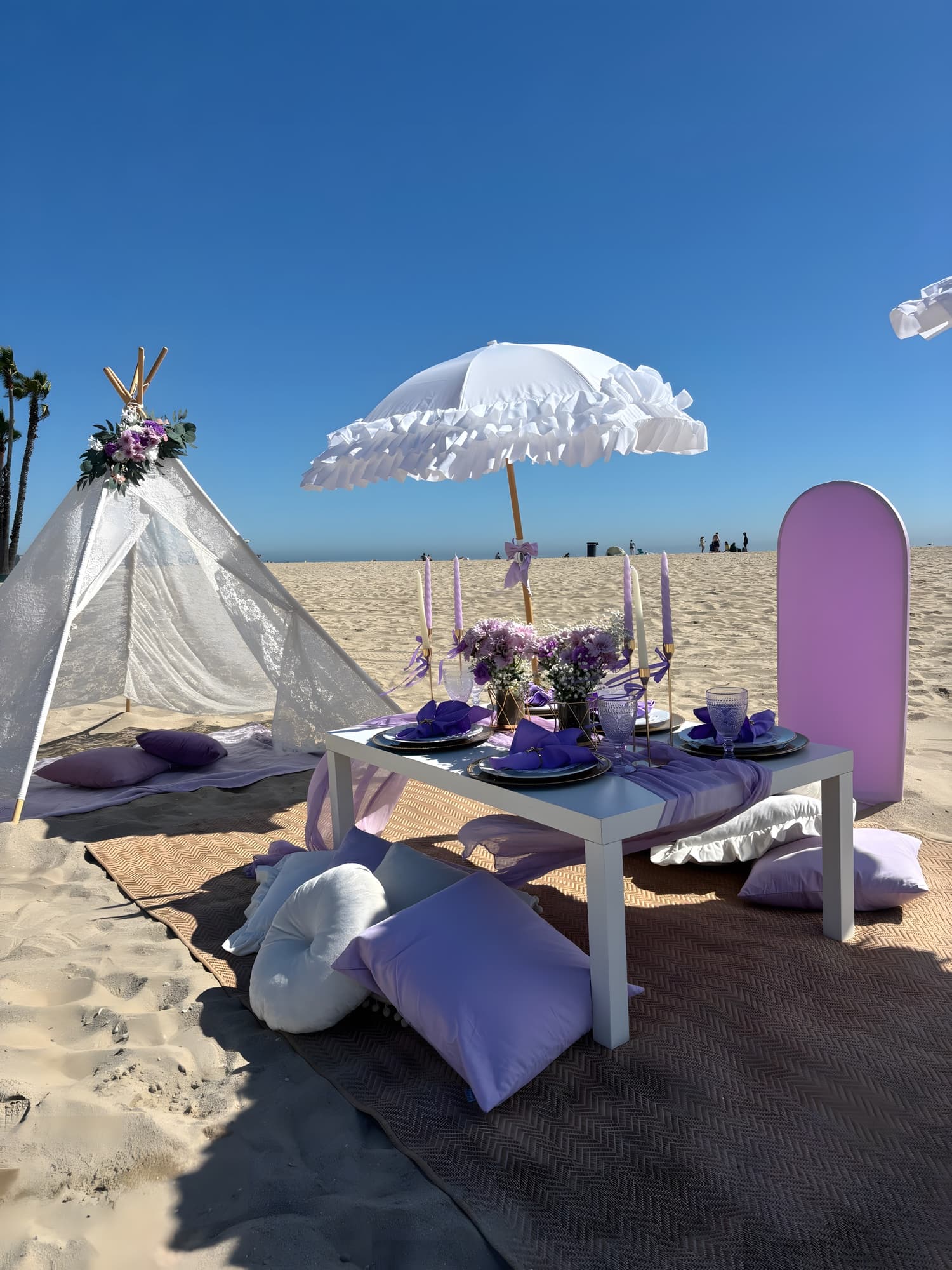 A lavender beach picnic with purple napkins, a ruffled umbrella and a lace teepee under a clear blue sky