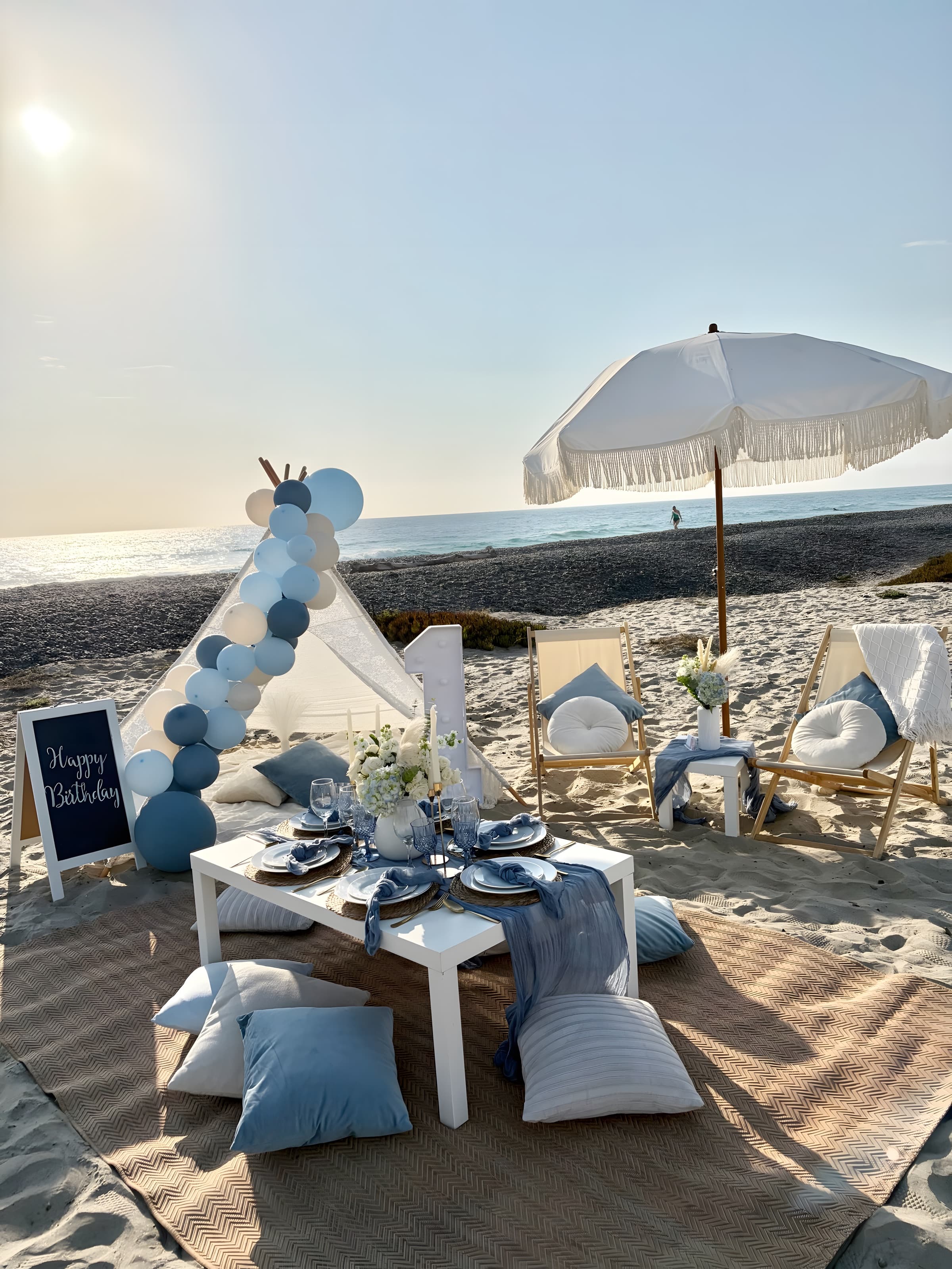 A dusty-blue first-birthday beach setup with a balloon-garland teepee, marquee “1” and a set table at sunset